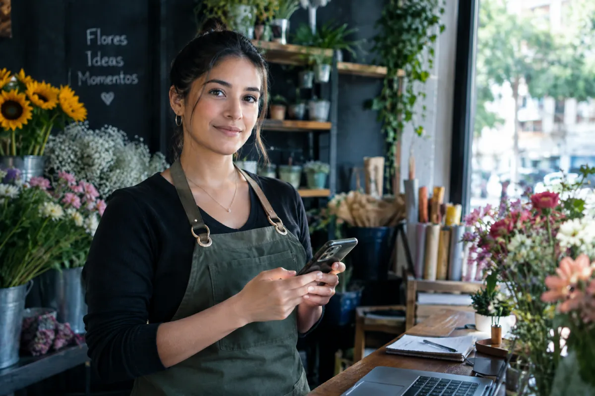 Florista sonriendo con su celular en la mano dentro de su tienda